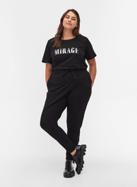 Femme portant un t-shirt noir "MIRAGE", un pantalon noir à cordon de serrage et des chaussures montantes noires à lacets, debout avec une jambe croisée.