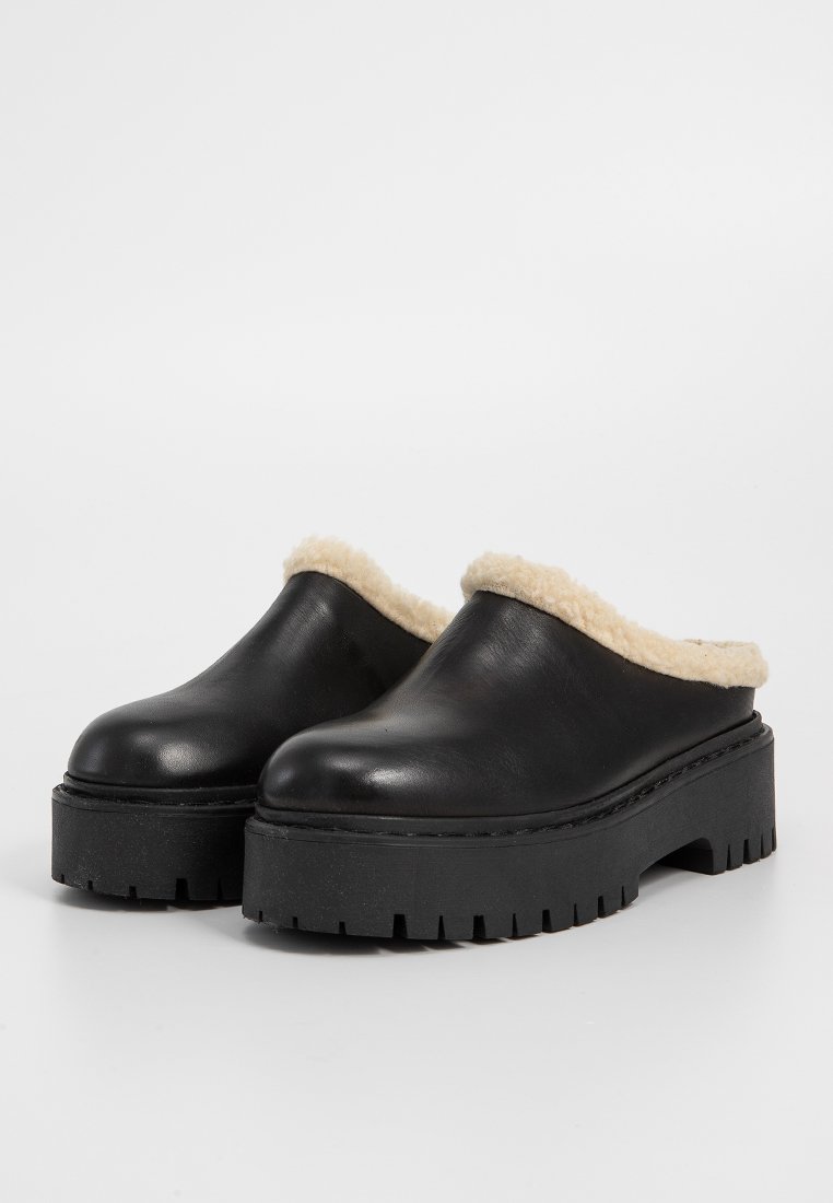 Sabots en cuir noir avec une doublure beige moelleuse et une épaisse semelle de plateforme noire texturée. Design à bout rond. Pas de matériel visible.