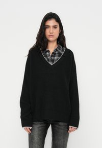 Melns pārāk lielizmēra adīts džemperis ar V-veida izgriezumu un kontrastējošu apakšmalu. Valkāts virs rūtainas pogas kreklā. Ir garas piedurknes un teksturēts audums.