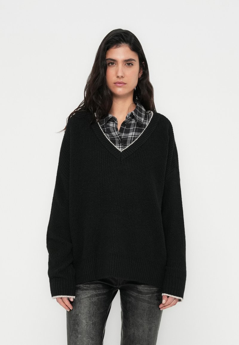 Melns pārāk lielizmēra adīts džemperis ar V-veida izgriezumu un kontrastējošu apakšmalu. Valkāts virs rūtainas pogas kreklā. Ir garas piedurknes un teksturēts audums.