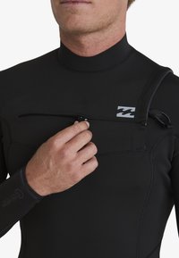 Traje de neopreno negro con alto cuello, que cuenta con un bolsillo con cremallera en el pecho y costuras en contraste. Incluye un logotipo en el lado izquierdo.