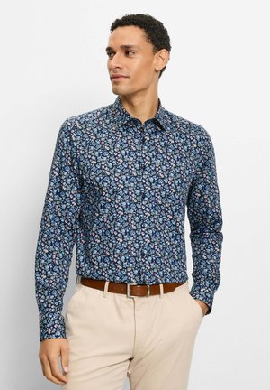 Mann trägt ein dunkelblaues, florales Hemd mit langen Ärmeln, in beige Hosen mit braunem Gürtel gesteckt, posiert mit einer Hand in der Hosentasche und schaut nach links.