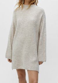 Femme portant une robe pull longue en tricot épais beige avec des manches longues sur un fond blanc uni.