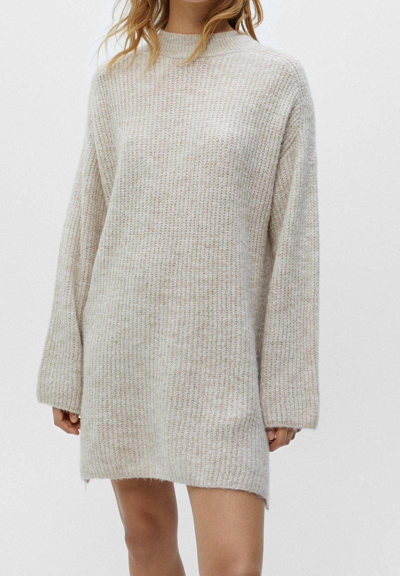 Femme portant une robe pull longue en tricot épais beige avec des manches longues sur un fond blanc uni.