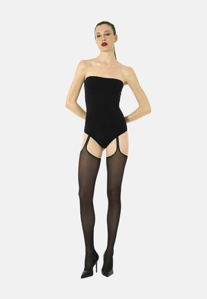 Donna con capelli legati indossa un body nero senza spalline, calze nere trasparenti con reggicalze e tacchi alti neri su sfondo uniforme.