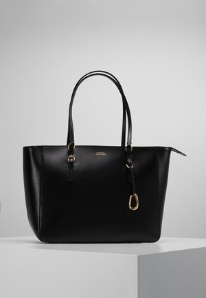 Black leather tote bag with double handles, gold hardware, and small front logo on a white pedestal against a gray background.