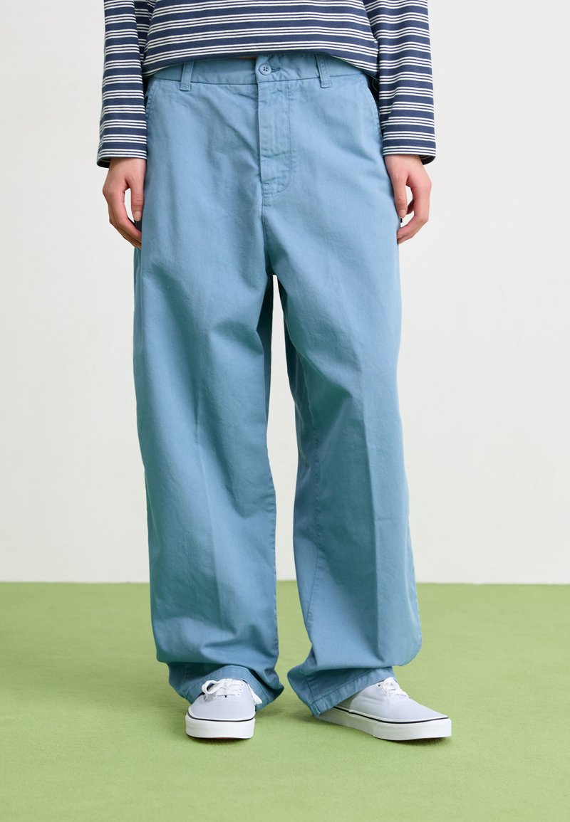 Light blue, wide-leg trousers feature a smooth texture and front button closure, paired with white sneakers on a green surface.