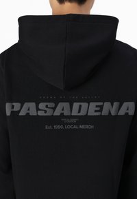 Crna dukserica s velikim sivim tekstualnim grafičkim natpisom na leđima koji glasi "PASADENA" s natpisom "CROWN OF THE VALLEY" iznad i "Est. 1990, LOCAL MERCH" ispod.
