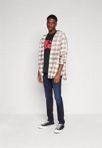 Chemise à carreaux beige et marron sur un t-shirt graphique noir. Un jeans bleu foncé et des baskets noires à semelles blanches complètent le look.