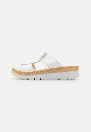 Weiße Sandale mit zwei ausgeschnittenen Riemen und einer strukturierten beigen Plattformsohle. Die Seitenansicht zeigt eine niedrige Wedge-Form und Details der gepolsterten Fußbett-Polsterung.