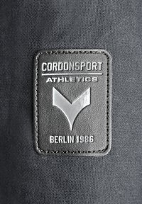 Schwarzer Stoff mit einem rechteckigen, genähten Lederpatch, auf dem „CORDONSPORT ATHLETICS“ und „BERLIN 1986“ in erhabener Schrift sowie einem Logodesign zu sehen sind.
