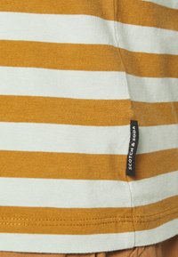 Stribet t-shirt i sennepsfarve og lyseblå, lavet af bomuld. Har en sort mærkat med "SCOTCH & SODA" på sidesømmen.