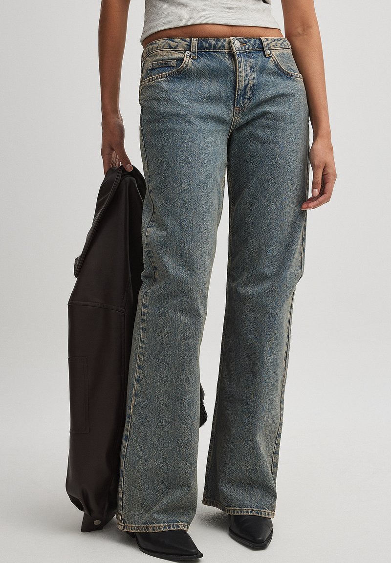 Vaqueros de denim azul claro con un corte relajado, que presentan una cintura de altura media, costuras visibles y un diseño clásico de cinco bolsillos. También hay calzado negro y una chaqueta oscura.
