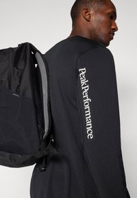 Sort rygsæk med et geometrisk mønster, justerbare stropper, grå mesh-bagside, og "Peak Performance" logo på langærmet skjorte.