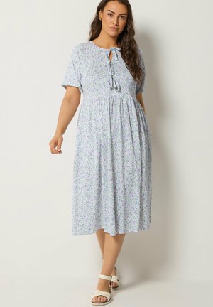 Kobieta w jasnofioletowej kwiecistej sukience midi z krótkimi rękawami i wiązaniem z przodu, ubrana w białe sandały na paski, stojąca na białym tle.