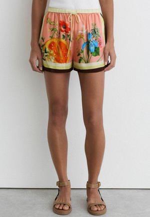Femme portant un short rose à cordon avec un imprimé floral et fruité coloré, associé à des sandales beiges à bride cheville, debout contre un mur blanc.
