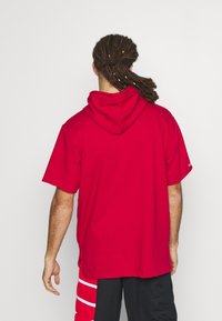 Persona con dreadlocks largos que lleva una sudadera corta roja y pantalones cortos negros con rayas rojas y blancas, de espaldas frente a un fondo simple.
