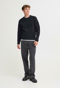 Svart crewneck genser lagd over en hvit skjorte, kombinert med mørkegrå jeans og svarte klumpete sko. Uformell passform med lange ermer.