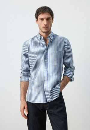 Uomo con capelli corti e scuri che indossa una camicia a righe verticali blu e bianche con bottoni e pantaloni scuri, in piedi con una mano in tasca.