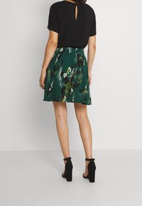 Schwarzes kurzärmliges Oberteil mit einem Schlüssel-augenrücken, kombiniert mit einem knielangen grünen Blütenrock, der abstrakte Muster aufweist und eine lockere Passform hat.