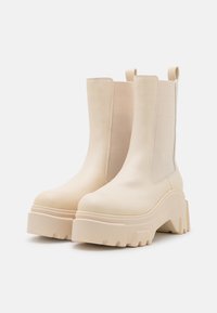 Une paire de bottes à plateforme épaisses, de couleur crème, avec des panneaux latéraux élastiques et des languettes de tirage, présentées sur un fond blanc.