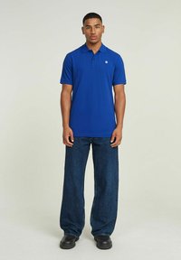 Polo blu con colletto, maniche corte e un piccolo logo, abbinato a jeans in denim scuro a vestibilità rilassata e scarpe nere.