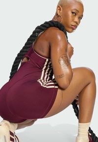 Athletic person in maroon sportswear and sneakers holds thick black battle rope, squatting and looking over shoulder against plain background.