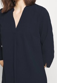 Mörkblå blus med V-ringning, halv ärm och bröstficka. Mjukt tyg med en slät yta och minimalistisk design.