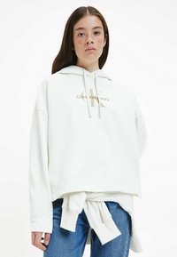 Felpa bianca con logo dorato "Calvin Klein Jeans". Presenta un cappuccio, una vestibilità oversize e un dettaglio legato in vita. Jeans in denim visibili sotto.
