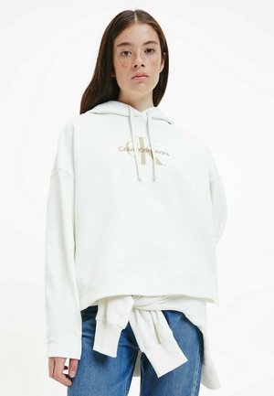 Felpa bianca con logo dorato "Calvin Klein Jeans". Presenta un cappuccio, una vestibilità oversize e un dettaglio legato in vita. Jeans in denim visibili sotto.