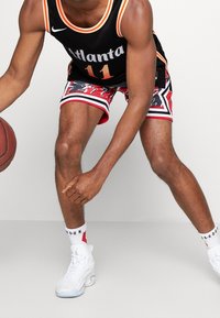 Camiseta de baloncesto negra con acentos naranjas, que presenta el texto "Atlanta" y el número 11. Pantalones cortos rojos con un patrón gráfico y ribete blanco. Zapatillas blancas.