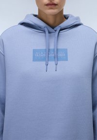 Lyseblå hettegenser med logo på framsiden, snøring og et mykt, strukturert stoff.