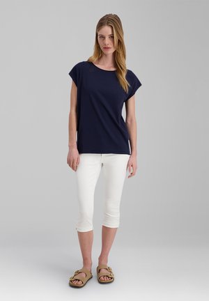 Young woman with long blonde hair wearing a navy blue short-sleeve top, white cropped pants, and tan sandals standing against a gray background.