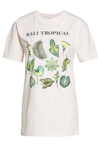 Gaiši bēšs kokvilnas t-krekls ar botānisko apdruku, kas attēlo dažādas zaļas lapas, virs dizaina melnā krāsā ir teksts "BALI TROPICAL".