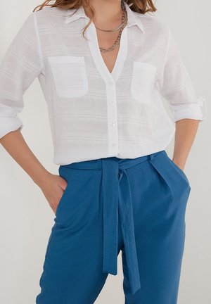 Woman wearing a semi-sheer white button-up shirt with pockets and rolled sleeves, paired with high-waisted blue pants tied at the waist.