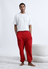 Γκρι μπλουζάκι με λογότυπο, κόκκινο παντελόνι φόρμας, χαλαρή εφαρμογή, μέση με ρυθμιζόμενο κορδόνι, στέκεται πάνω σε ανοιχτό γκρι χαλί κοντά σε υφασμάτινο καναπέ ανοιχτού χρώματος.