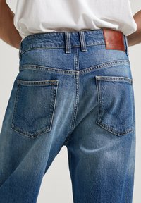Modré džíny z denimu so vyblednutým povrchom, so zošitým koženým zadným panelom v hnedej farbe a dvomi zadnými vreckami s dekoratívnym šitím.