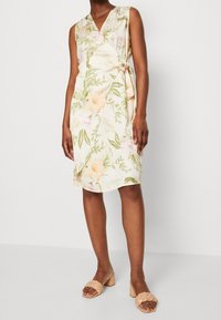 Mouwloze jurk met bloemenpatroon in roomkleur, met groene bladeren en zachte roze bloemen. Wikkelontwerp met een zijtasje, van lichtgewicht stof.