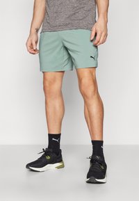 Light green athletic shorts with an elastic waistband, paired with black sneakers and socks featuring a white logo. Gray shirt above the waist.