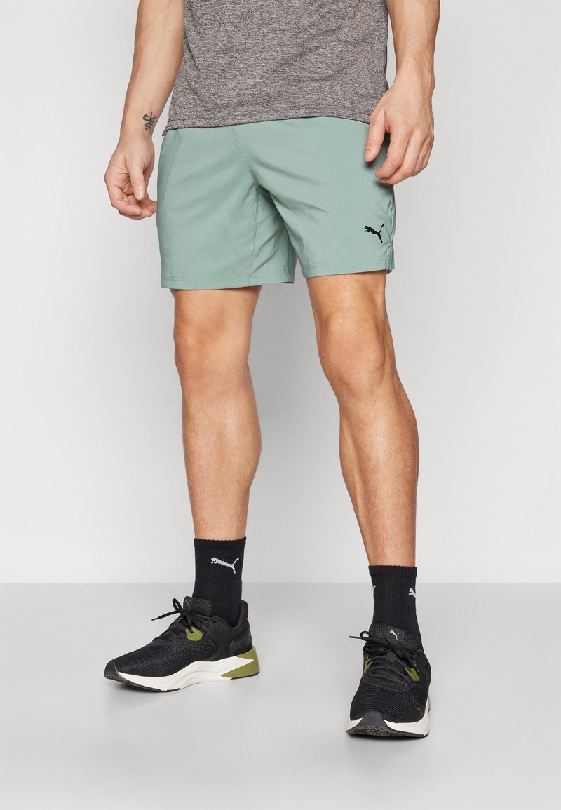 Light green athletic shorts with an elastic waistband, paired with black sneakers and socks featuring a white logo. Gray shirt above the waist.