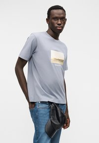 Ανοιχτό γκρι βαμβακερό μπλουζάκι με ορθογώνιο γραφικό τύπωμα προκυμαίας. Συνδυάζεται με μπλε τζιν και μαύρη τσάντα, τοποθετημένη στη μέση.