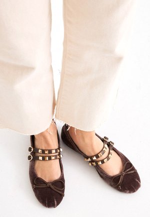 Bailarinas de terciopelo marrón con detalle de lazo, diseño de dos tiras con acentos metálicos, combinadas con pantalones claros desgastados.