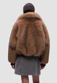 Giacca di pelliccia marrone con ampio colletto, texture soffice e polsini a costine, abbinata a una gonna grigia, vista da dietro.