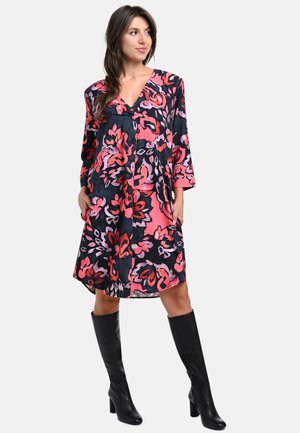 Florales Kleid in Schwarz mit pinken und roten Mustern, ausgestattet mit einem V-Ausschnitt, drei Viertelärmel und einer lockeren Passform. Kombiniert mit schwarzen kniehohen Stiefeln.