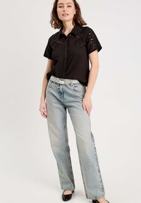 Chemise noire à manches courtes avec des détails en œillets, associée à un jean large en denim délavé et une ceinture en argent, sur un fond uni.