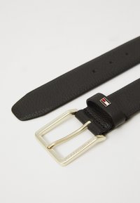 Ceinture en cuir noir texturé avec une boucle rectangulaire dorée gravée du nom de la marque et un petit logo multicolore sur l'anneau de la ceinture.