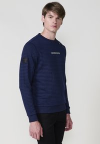 Joven con pelo castaño claro viste una sudadera de textura azul marino con el logo "KOROSHI" y pantalones negros, de pie frente a un fondo blanco.
