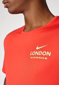 Κόκκινο αθλητικό μπλουζάκι από ελαφρύ ύφασμα με κοψίματα στο λαιμό, υφασμάτινη επιφάνεια και εκτυπωμένο κείμενο: "ΛΟΝΔΙΝΟ" και λεπτομέρειες απόστασης.