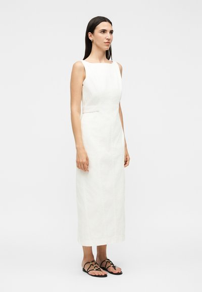 Γυναίκα στέκεται φορώντας ένα λευκό αμάνικο φόρεμα μέχρι τους αστραγάλους και μαύρα σανδάλια με λουριά και φλατ πάτο, σε απλό φωτεινό φόντο.