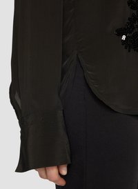 Zwarte satijnen blouse met een glanzende textuur, heeft een gestructureerde mouw en is versierd met zwarte kralen. Zijslits detail nabij de zoom.